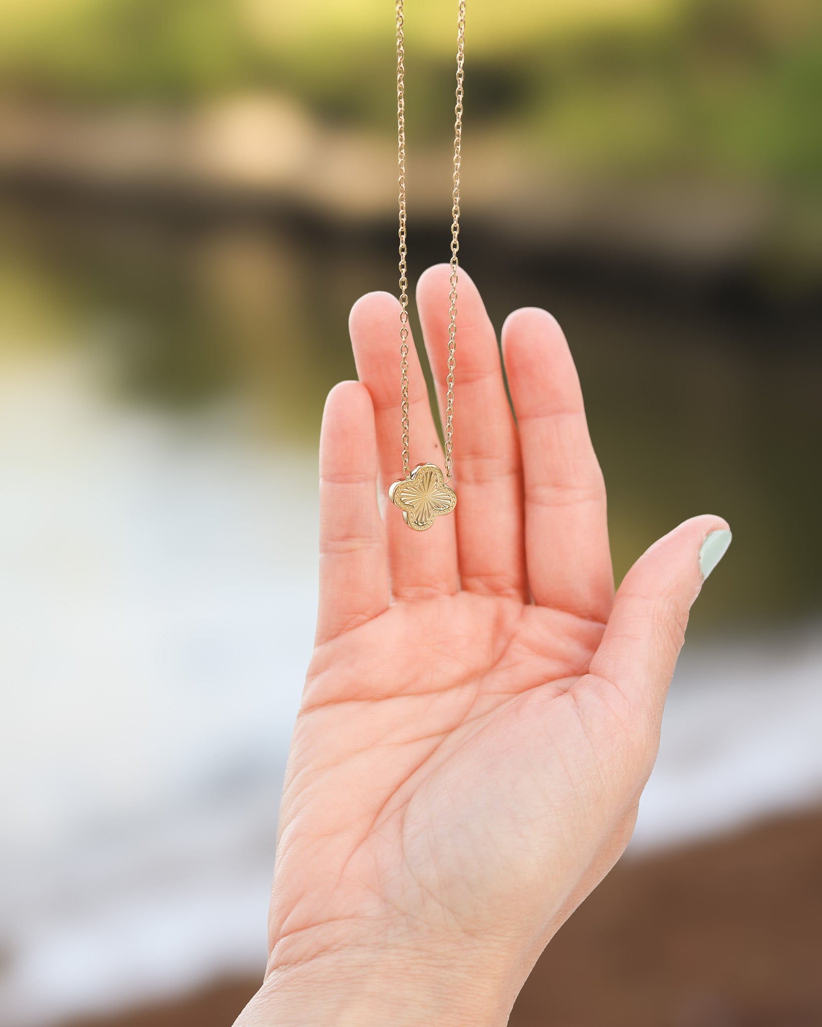Delicate gold clover necklace hanging from fingers with blurred background
