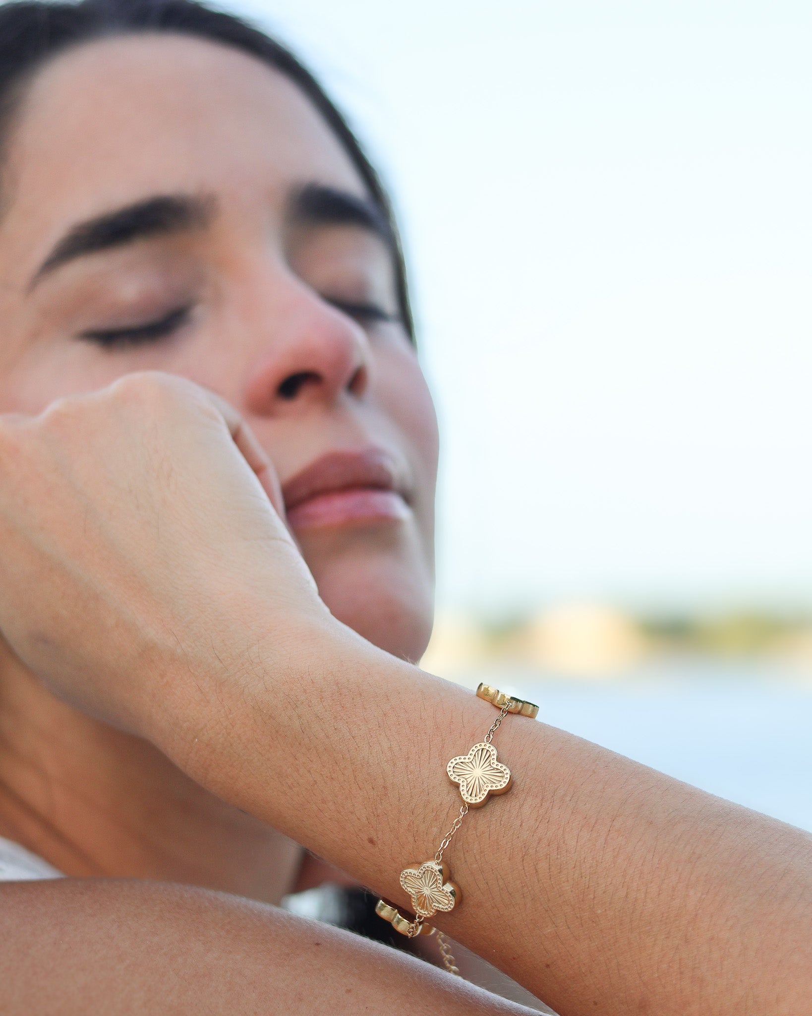 Woman wearing a gold bracelet with floral designs by a body of water.