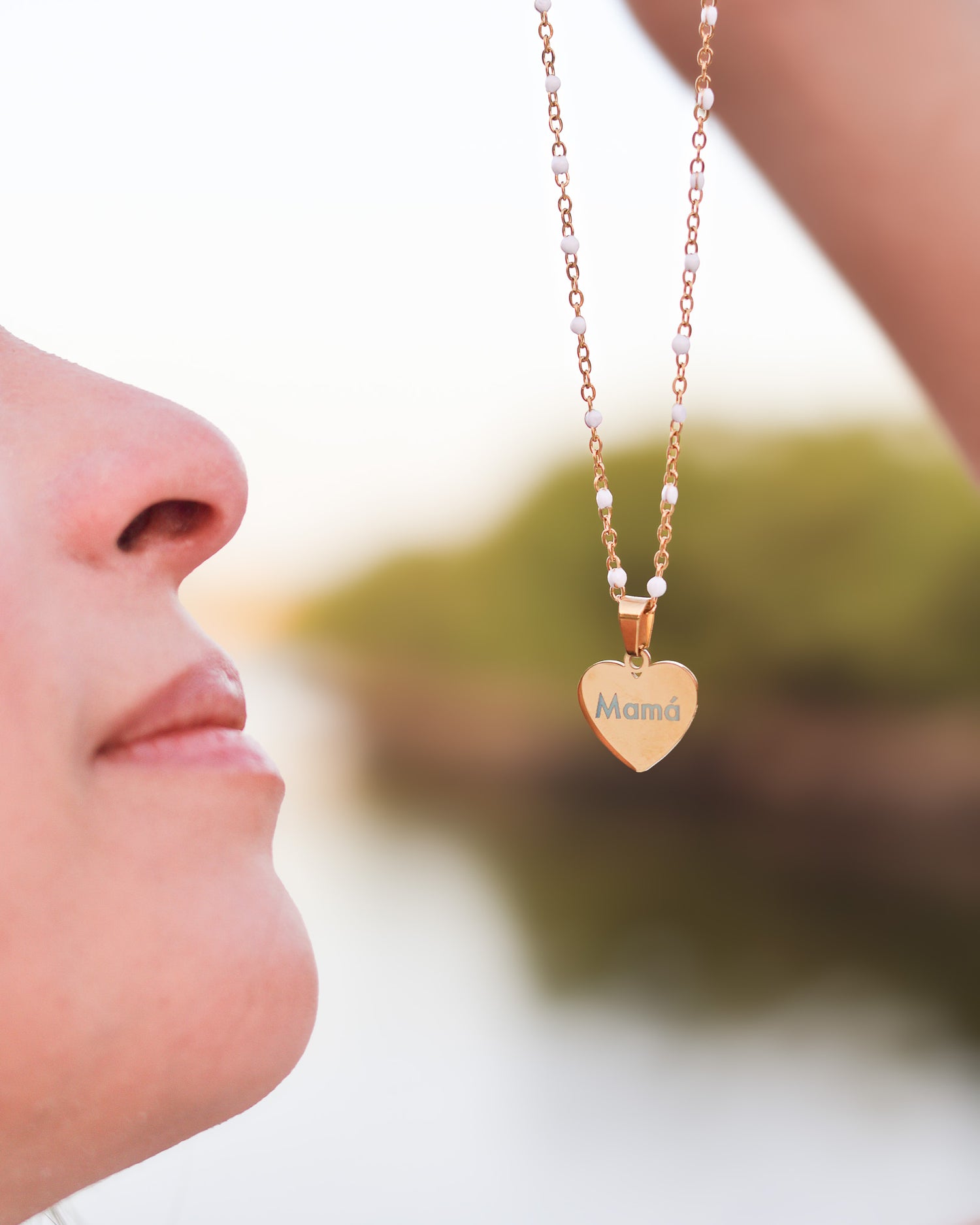 Gold heart-shaped necklace with custom engraving, placed on soft beach sand near white beads
